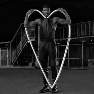 Black and white portrait of a man standing outdoors holding a large heart-shaped metal frame in front of him.