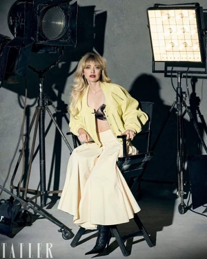Imogen Poots wearing a pale yellow jacket and cream wide-leg trousers seated in a director's chair between studio lights.