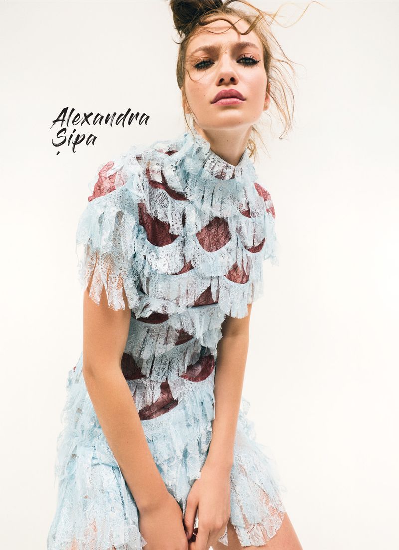 Young fair-skinned woman poses wearing a light blue and maroon lace dress in a bright, clean photo studio setting.