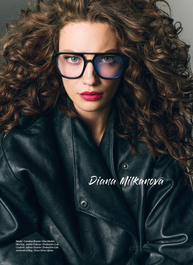 Close-up portrait of a brunette woman with curly hair wearing glasses and a black leather jacket in a studio setting with soft lighting.