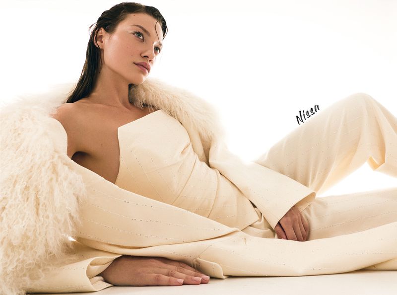 A woman in a cream-colored pantsuit with a faux fur shawl lies on a white surface in a fashion-style pose, looking slightly away from the camera.