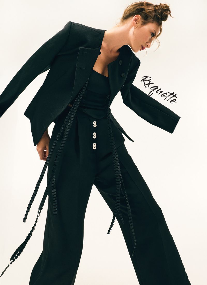 Fashion shot of a woman in a black blazer, pants, and top, angled pose against a white backdrop, showcasing modern design and style.