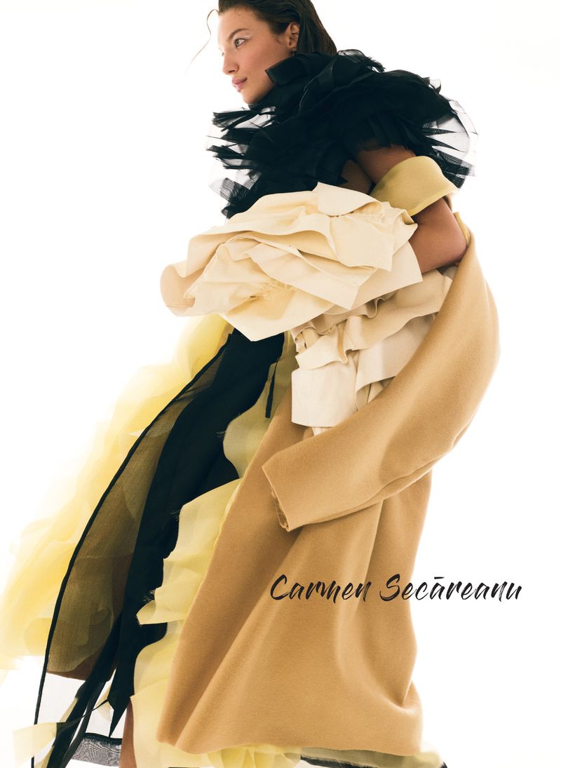 Editorial fashion shot features a woman with light skin and long hair wearing a tan coat and black and cream-colored tulle garment in studio.