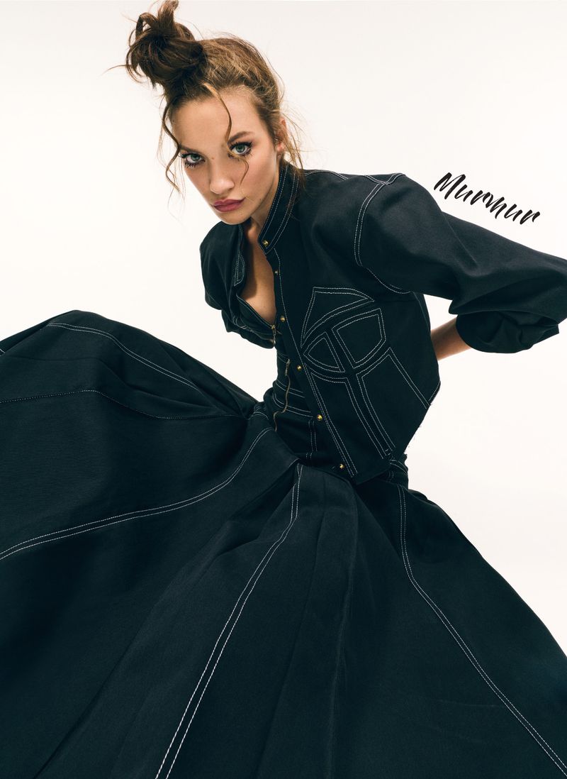 A woman poses in a black couture dress with white trim, captured in a high-angle shot against a bright backdrop, showcasing fashion and elegance.