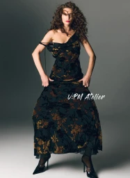 Model with curly brown hair wearing a dark floral dress and black heels against a grey studio backdrop.