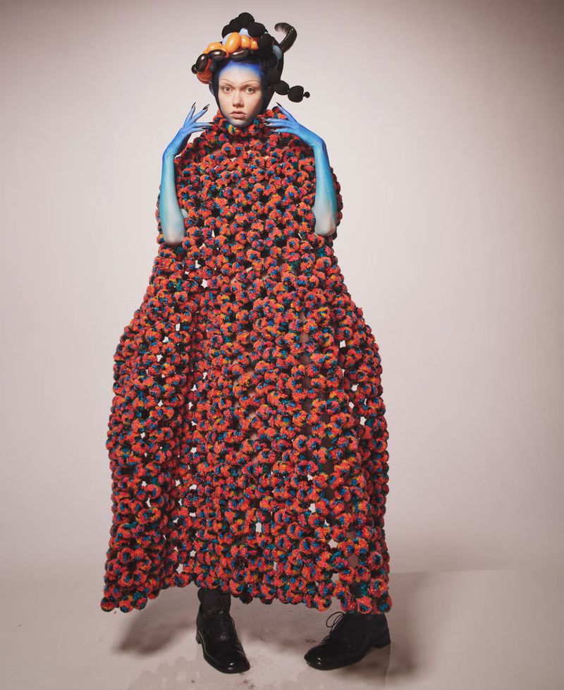 Conceptual fashion portrait of a person with blue skin and ornate headdress wearing a large, textured red and black cloak in studio setting.