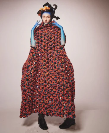 Conceptual fashion portrait of a person with blue skin and ornate headdress wearing a large, textured red and black cloak in studio setting.