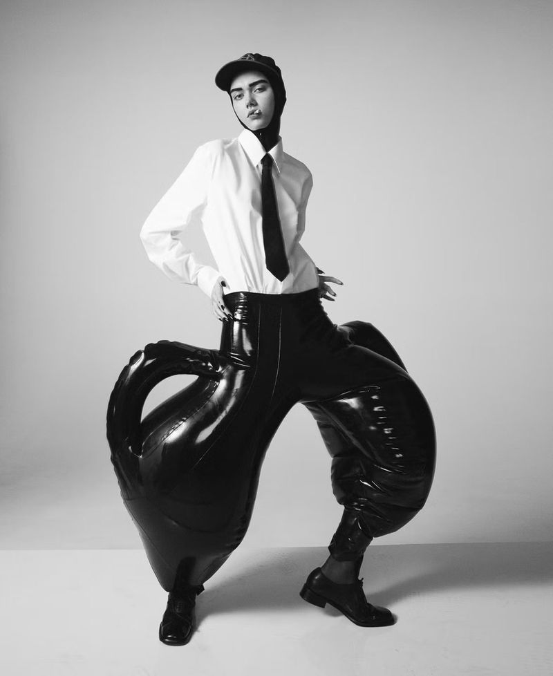 Monochrome fashion portrait of a person in an oversized shiny black outfit, white shirt, tie, and cap against a plain background.