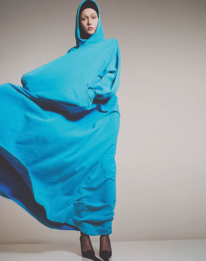 Fashion shot of a woman posing in a hooded blue dress with black stocking feet in a studio against a light grey background.