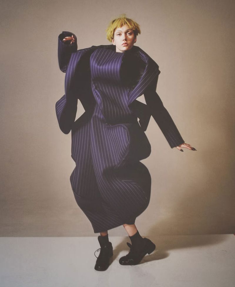 Fashion photography featuring a model in an oversized striped suit and black boots against a neutral backdrop in a full-length studio shot.