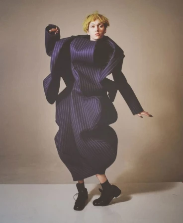 Fashion photography featuring a model in an oversized striped suit and black boots against a neutral backdrop in a full-length studio shot.