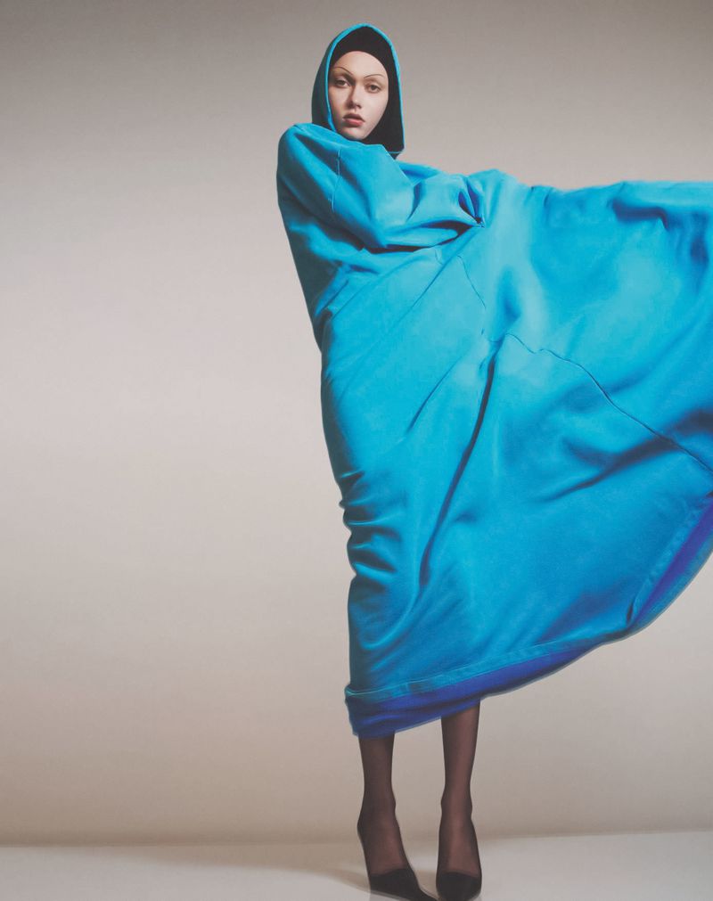 Model wearing an oversized bright blue hooded garment and black heels posing against a neutral studio backdrop.