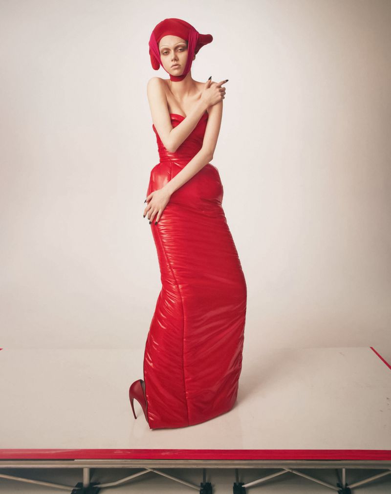 Model wearing a glossy red strapless gown and matching headwear against a white studio background