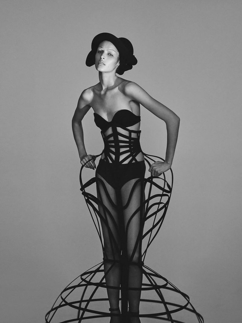 A model poses wearing an avant-garde black cage crinoline dress, structural corset, and sculptural hat against a grey seamless backdrop.