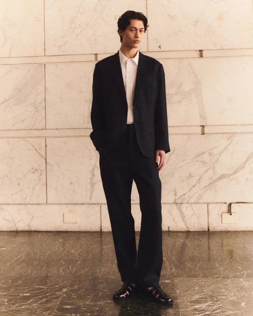 Full-length shot of a young adult male wearing a dark suit with a white shirt, standing in front of a light marble wall and floor.