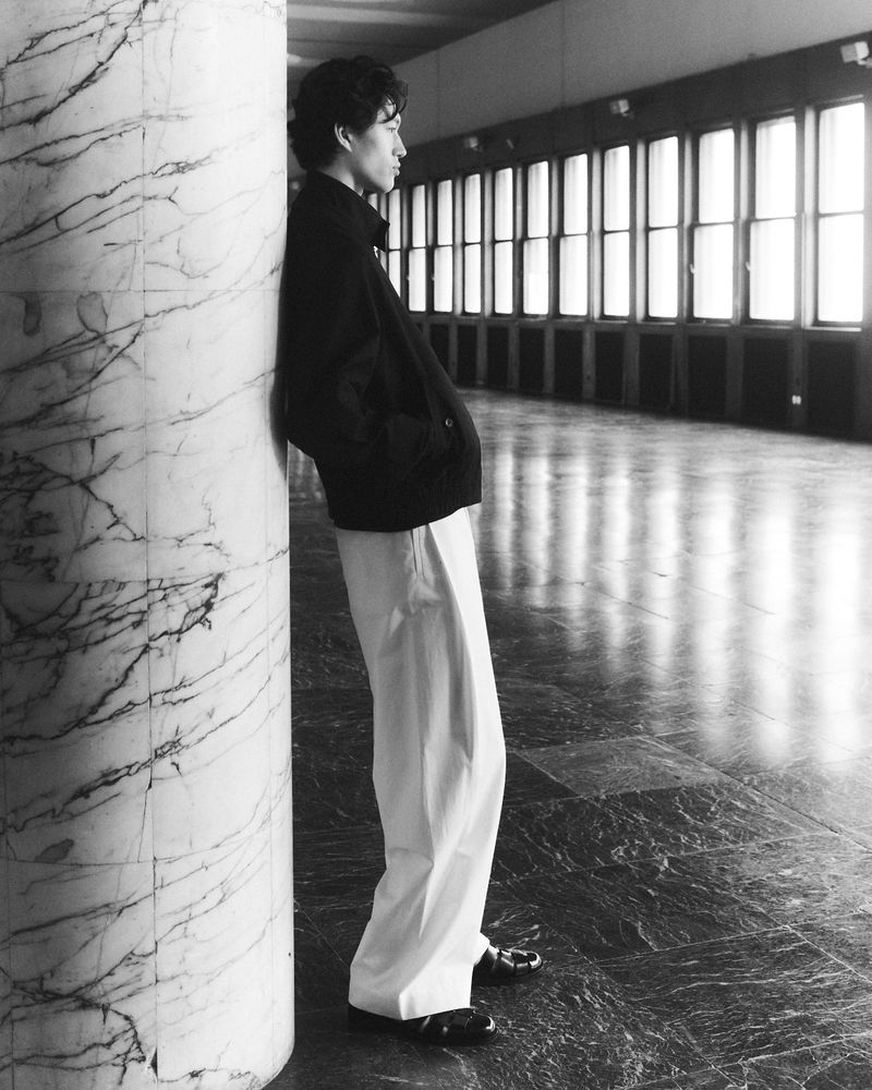 Black and white shot of an adult leaning against a marble column in a hallway with bright windows. Wearing loose white pants.