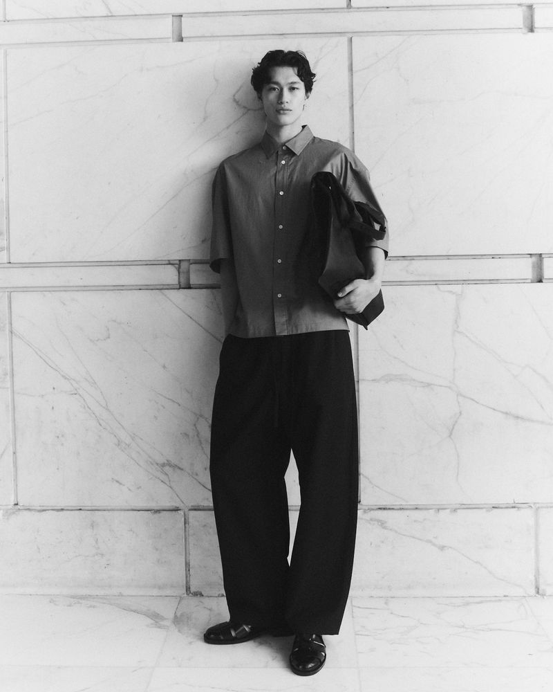 Monochrome portrait of a young man in a button-down shirt and wide-leg pants, holding a tote bag against a marble wall.
