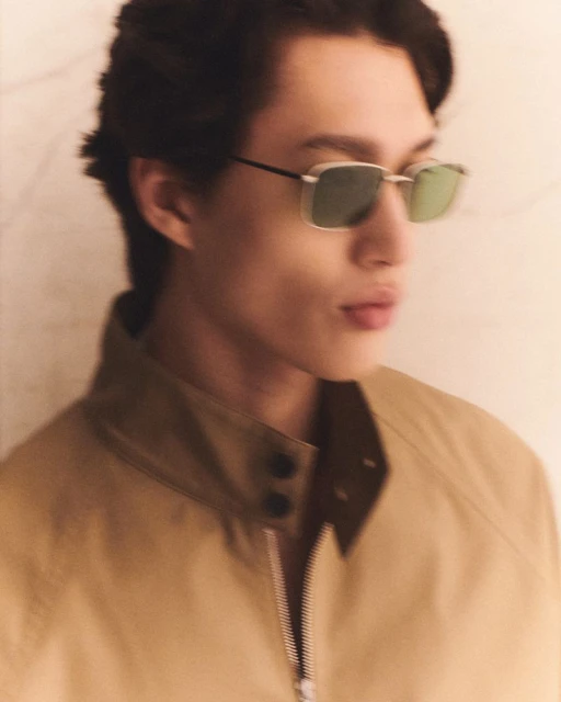Close-up portrait of a young adult male wearing square sunglasses and a tan-colored jacket with a zipper in a neutral indoor setting.