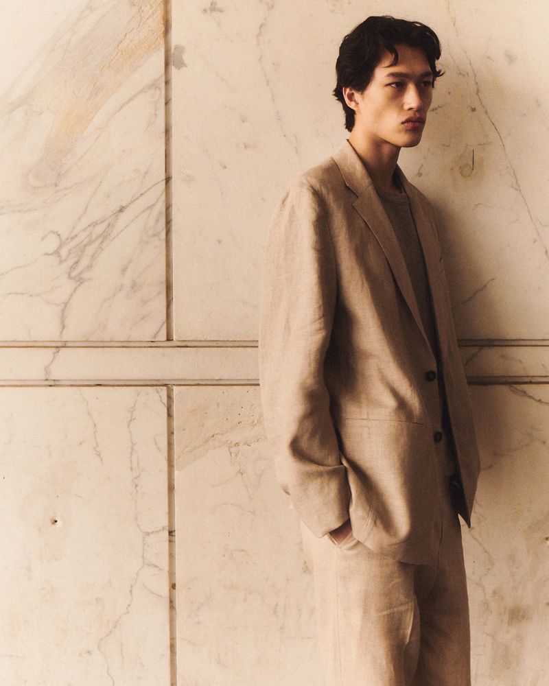 Model in a tan linen suit leaning against a veined marble wall for the COS Spring/Summer 25 Collection campaign.