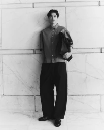 Man wearing a button-down shirt and loose trousers leaning against a large stone block wall, holding a dark bag.