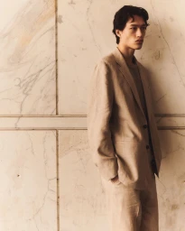 Model in a tan linen suit leaning against a veined marble wall for the COS Spring/Summer 25 Collection campaign.