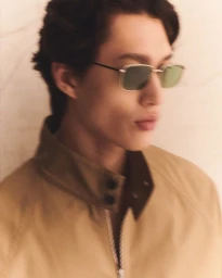 Model wearing tinted sunglasses and tan jacket against a pale textured background
