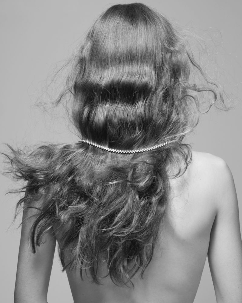 Black and white portrait of an adult female with wavy hair wearing a diamond necklace on her back in front of a neutral backdrop.