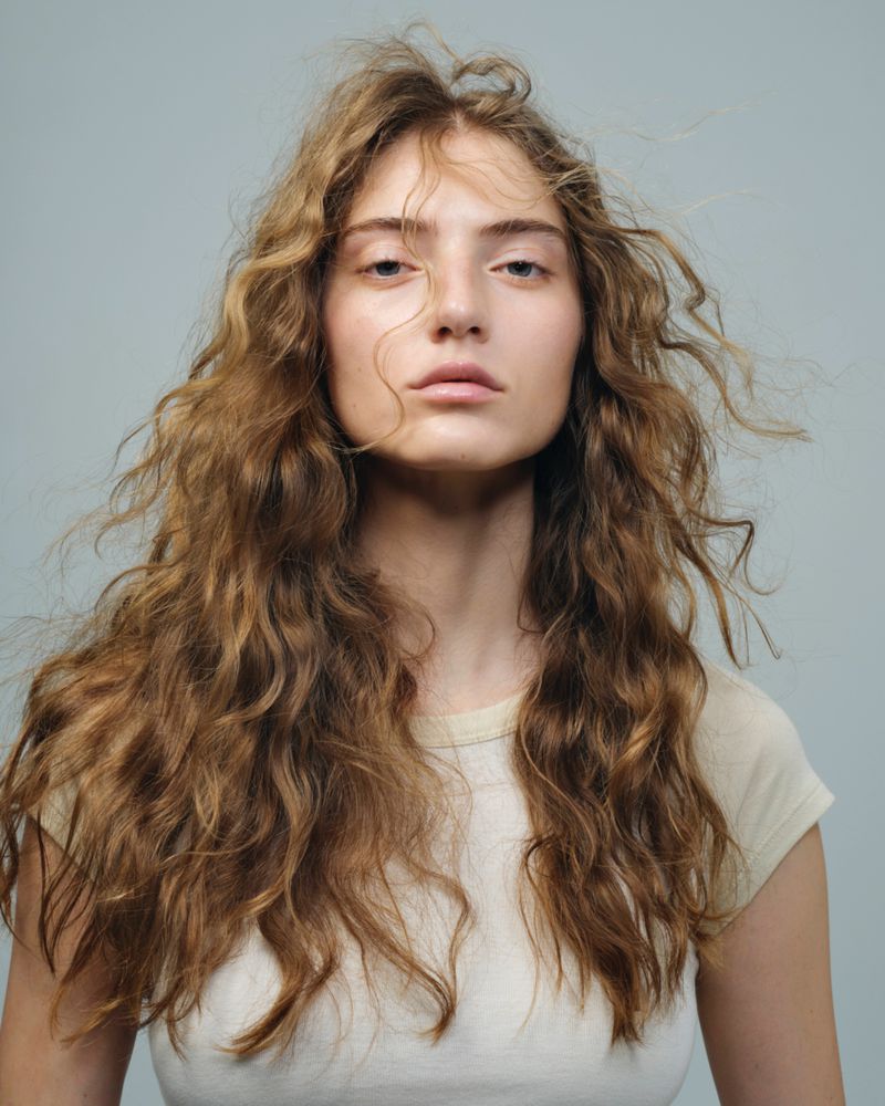 Portrait of a woman with long, wavy, light brown hair and neutral expression, wearing a cream-colored shirt, against a plain grey background.