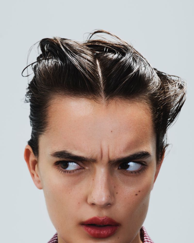 Close-up portrait of a young adult woman with dark hair styled back, looking to the side against a neutral background in a studio shot.