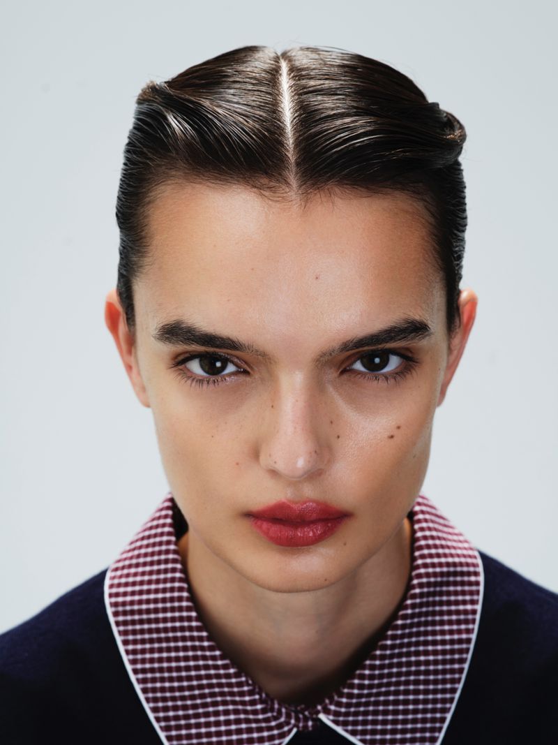 Close-up portrait of a young adult woman with slicked-back brunette hair, light skin, wearing a patterned collar and dark sweater.