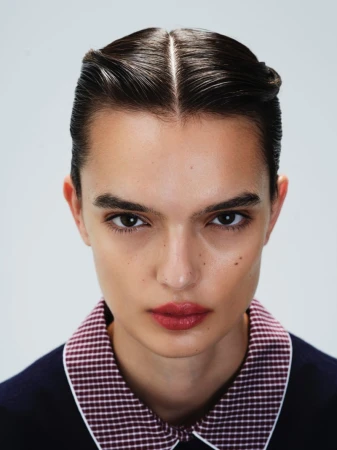 Close-up portrait of a young adult woman with slicked-back brunette hair, light skin, wearing a patterned collar and dark sweater.