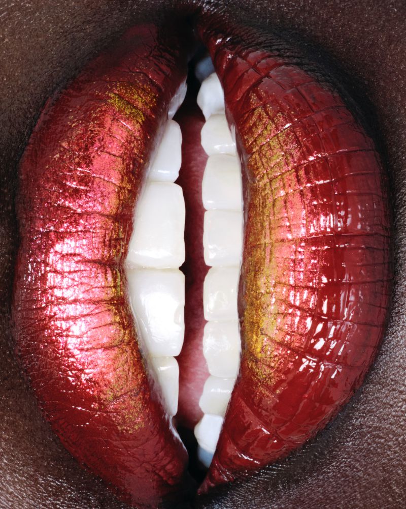 Close-up studio shot of a person's face showing teeth and glossy lips with red and gold lipstick against a dark skin background.