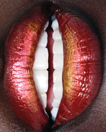 Close-up studio shot of a person's face showing teeth and glossy lips with red and gold lipstick against a dark skin background.