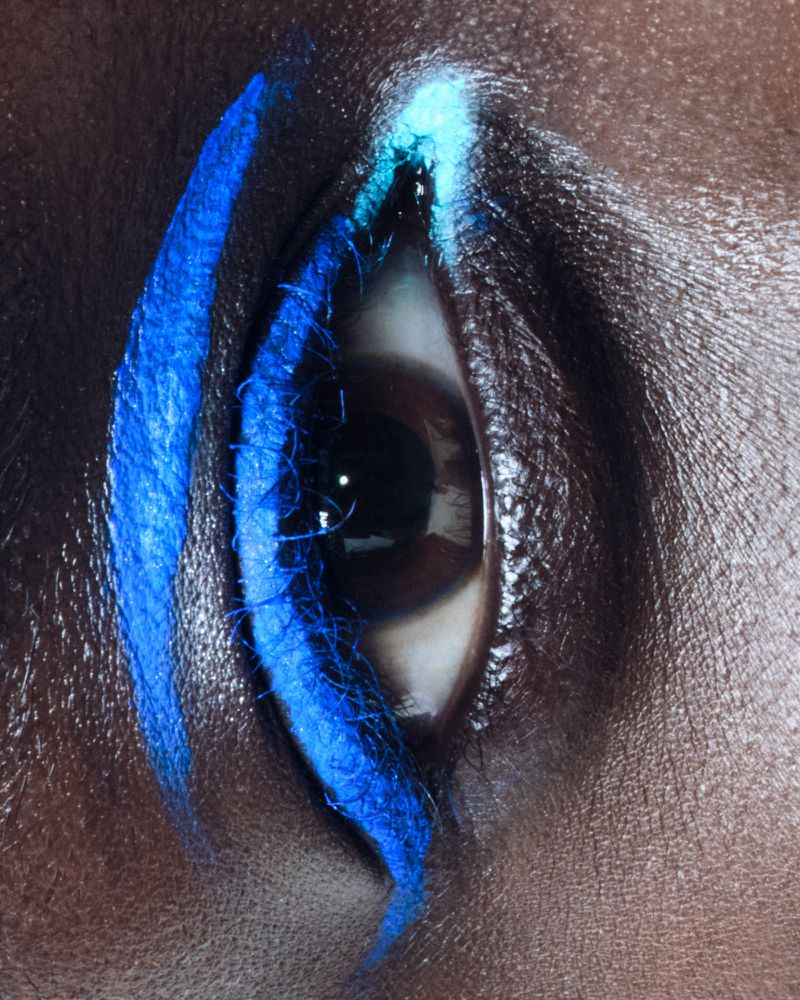 Close-up of a human eye with blue makeup. The makeup is applied around the eye. The person has dark skin and dark eyelashes.