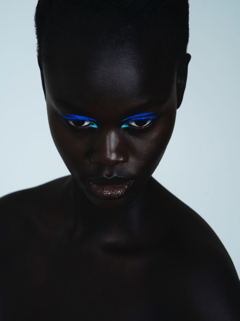 Close-up studio portrait of a Black woman with blue and turquoise eyeliner gazing downwards, set against a pale background.