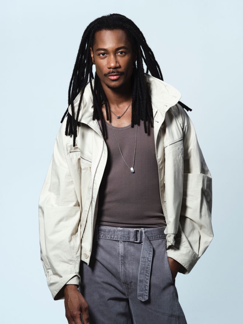 A man with dreadlocks poses in a light jacket, tank top, and pants against a bright background in a studio portrait.