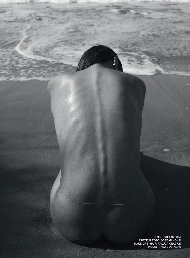 Monochrome photograph of a woman seated on a sandy beach facing the ocean waves, captured from behind focusing on her spine.