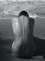 Monochrome photograph of a woman seated on a sandy beach facing the ocean waves, captured from behind focusing on her spine.