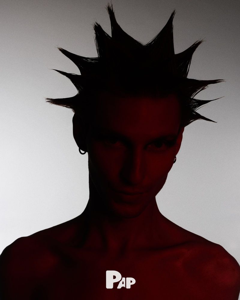 Red-toned portrait of a person with spiky hair, facing the camera with a neutral expression against a gradient light background.