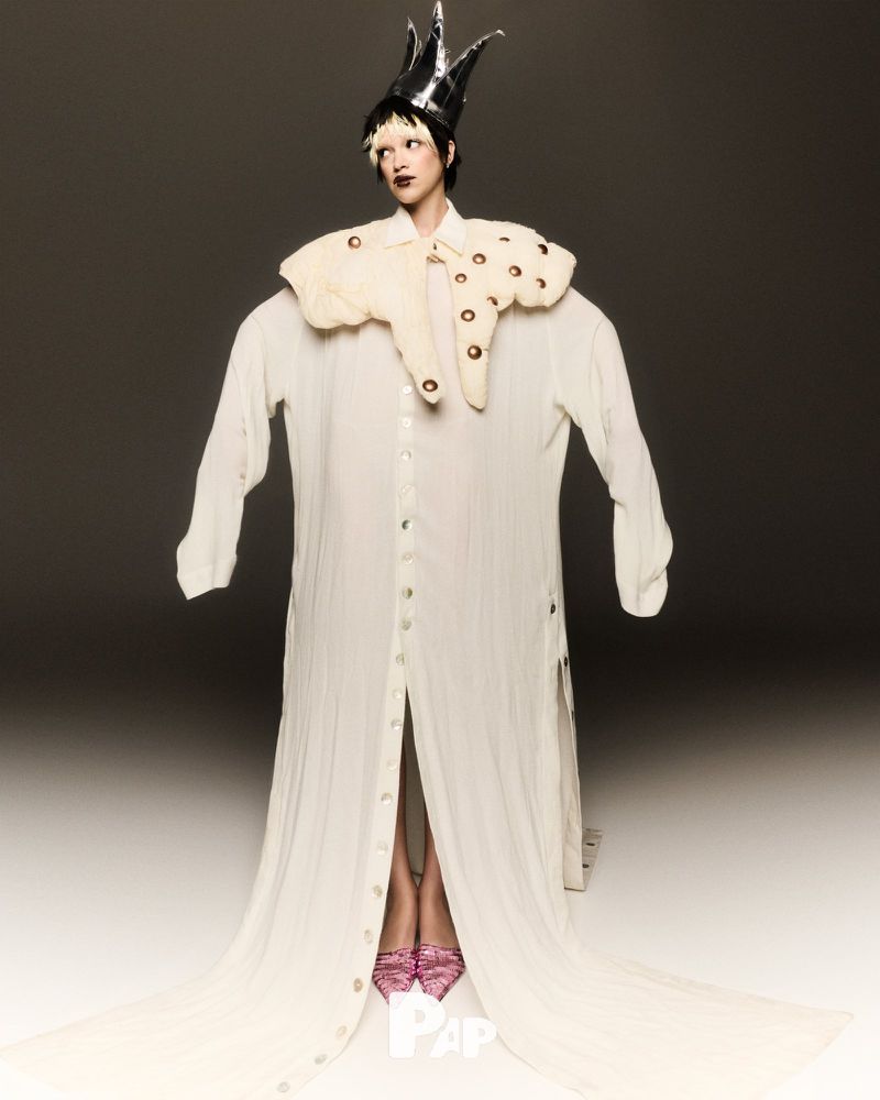 Adult female stands in a photo studio wearing an oversized white dress shirt, a ruffled collar with brown accents, a crown, and pink shoes.