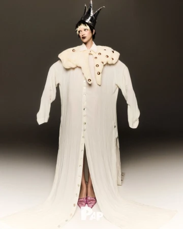 Adult female stands in a photo studio wearing an oversized white dress shirt, a ruffled collar with brown accents, a crown, and pink shoes.