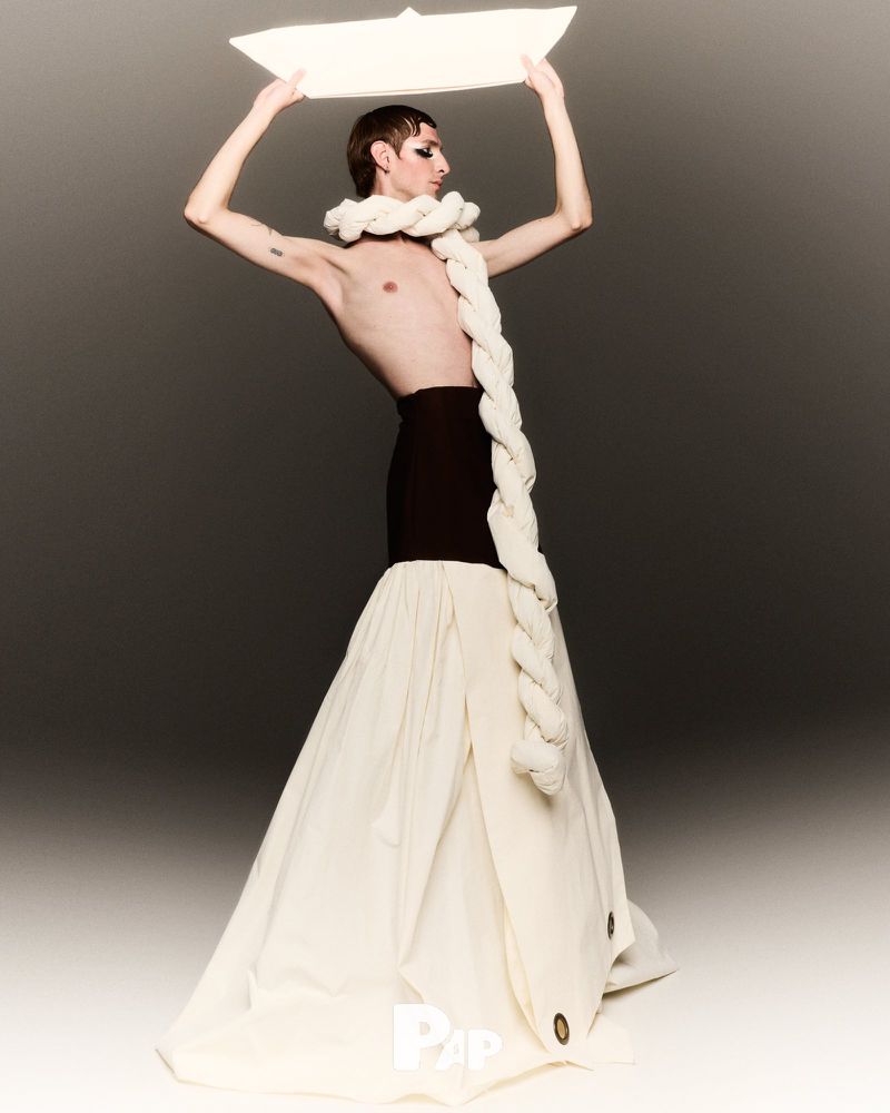 Fashionable individual poses in avant-garde clothing with white rope and black skirt holding paper boat. Light background.
