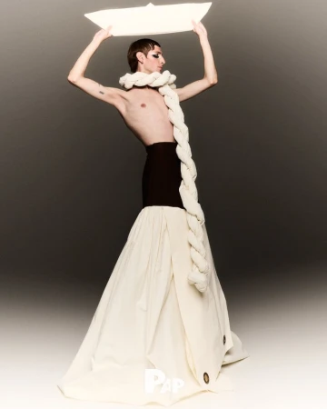 Fashionable individual poses in avant-garde clothing with white rope and black skirt holding paper boat. Light background.