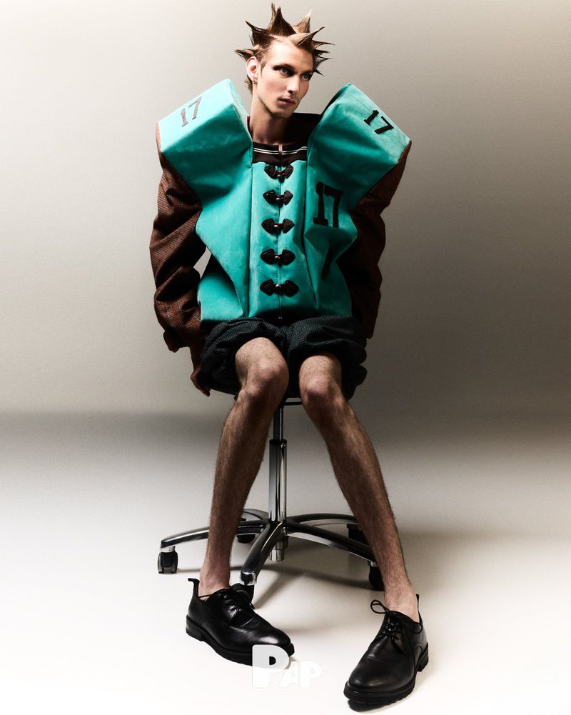 Male fashion model with spiky hair wearing a teal and brown padded vest sitting on a rolling chair in a photo studio.