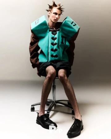 Male fashion model with spiky hair wearing a teal and brown padded vest sitting on a rolling chair in a photo studio. 