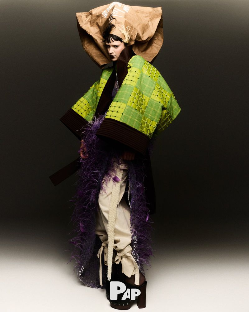 Avant-garde fashion portrait: a model in sculptural green jacket, oversized paper headpiece, and chunky platform shoes against a grey backdrop.