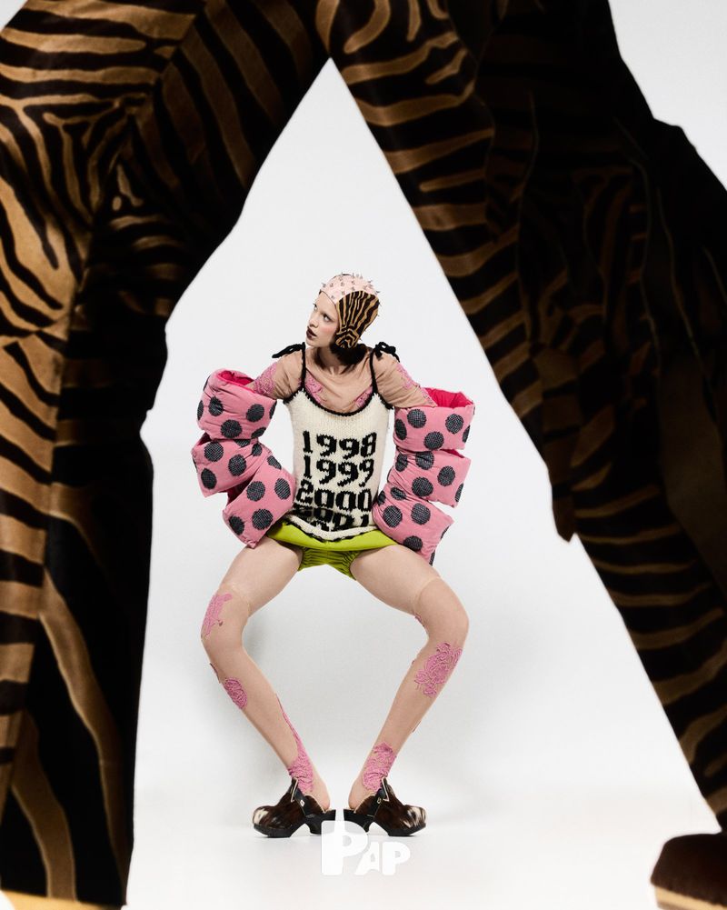 Eccentric fashion shoot: a person in unique attire, featuring bold patterns, polka dots, and distinctive headwear, stands against a white backdrop.