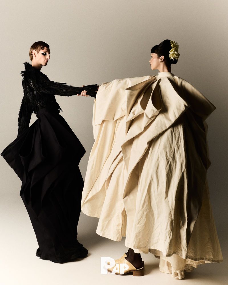 Two fashion models in sculptural dresses hold hands in a studio setting, one in black feathers and the other in draped cream fabric.