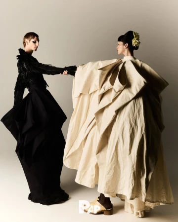 Two fashion models in sculptural dresses hold hands in a studio setting, one in black feathers and the other in draped cream fabric.