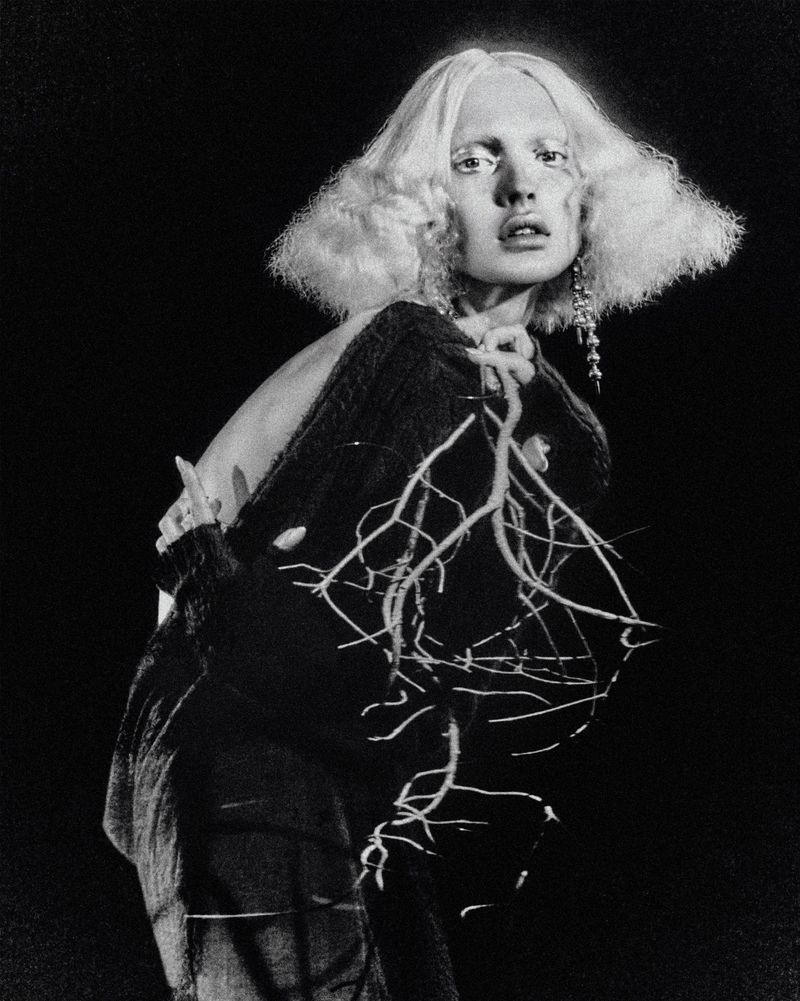 Monochrome portrait of a fashion model wearing a dark, embellished sweater against a dark background in a studio setting.